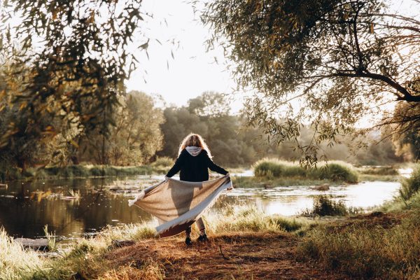 Back view of young woman with blanket in hands enjoying beautiful view on river with sunlights around. Happy female spending free time on nature.